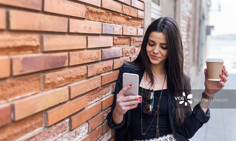 年轻漂亮的女人，靠在墙上，喝着外卖咖啡和智能手机图片素材