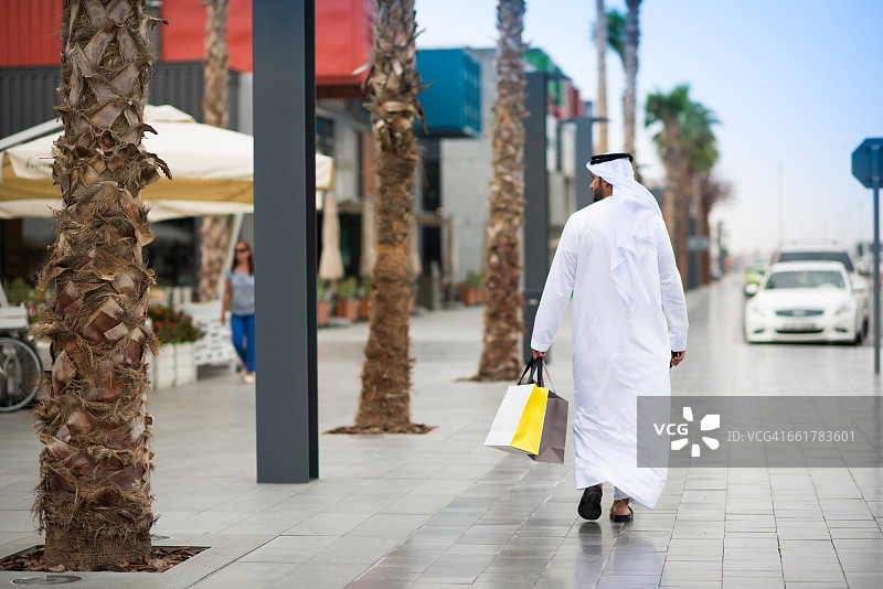 男子穿着dishdasha走在街上携带购物袋，迪拜，阿拉伯联合酋长国的后视图图片素材