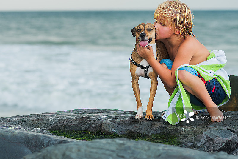 一个小男孩蹲在海滩的岩石上，抱着一只宠物狗图片素材