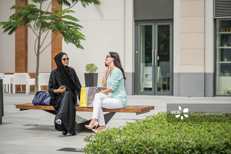 年轻的中东妇女穿着传统服装和女性朋友坐在长凳上，迪拜，阿拉伯联合酋长国图片素材