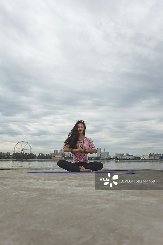 年轻女子练习瑜伽在祈祷位置对着河流在城市图片素材