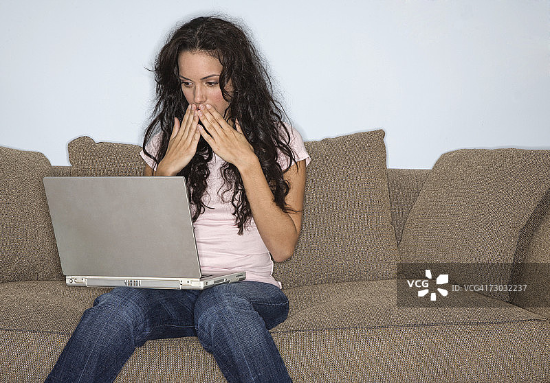 年轻女子坐在沙发上用手提电脑，用手捂着嘴图片素材