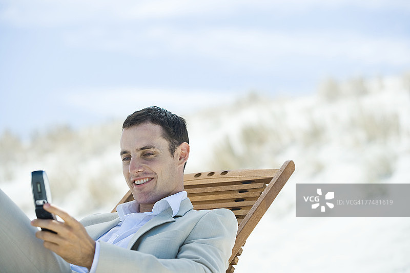 商人坐在沙滩的躺椅上，低头看着手机，微笑着图片素材