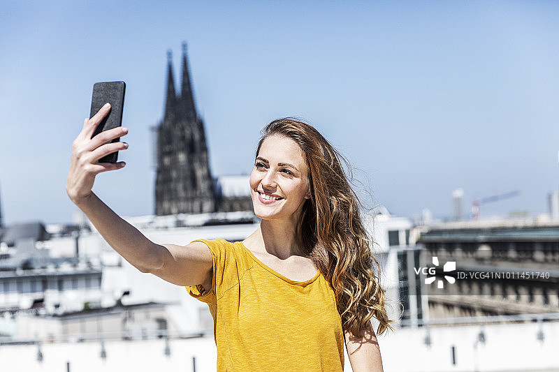 德国，科隆，一名微笑的女子在屋顶露台上用智能手机自拍图片素材