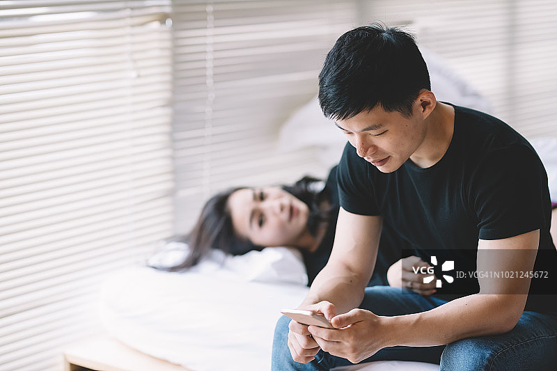 亚洲华人丈夫坐在床上发短信，妻子睡在他旁边图片素材