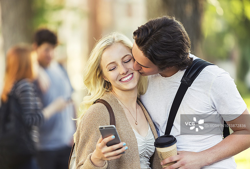 一名年轻女子在大学校园里用智能手机查看社交媒体时，一名年轻男子亲吻了她的脸颊图片素材