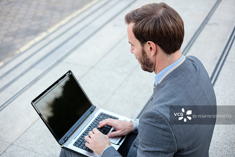 年轻英俊的商人在办公大楼前用笔记本电脑工作。高角度视图图片素材