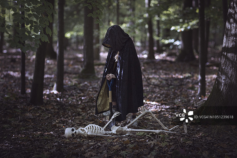 穿着兜帽黑色斗篷的孩子在黑暗的森林里用铲子挖一个洞来寻找一具假骨架图片素材