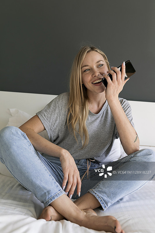 快乐的年轻女子坐在床上玩手机图片素材
