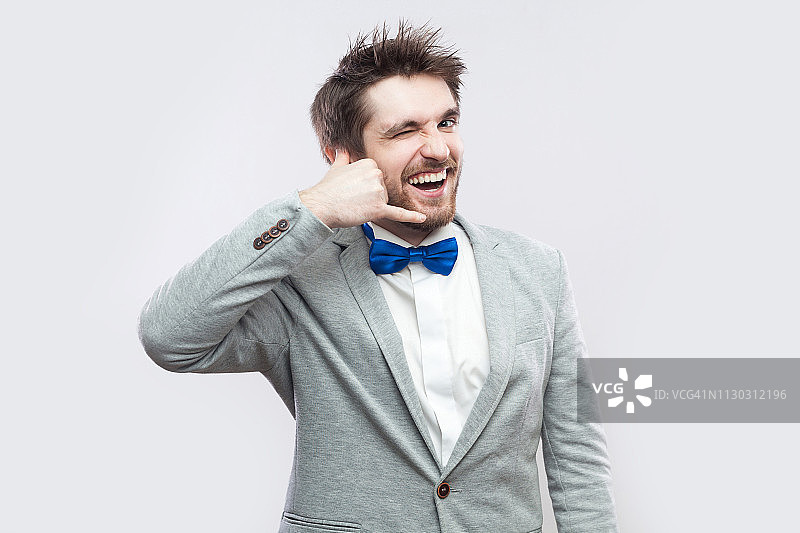 滑稽的眨眼快乐英俊的胡子男人的肖像在休闲灰色西装和蓝色领结与呼叫手势手和看相机。图片素材