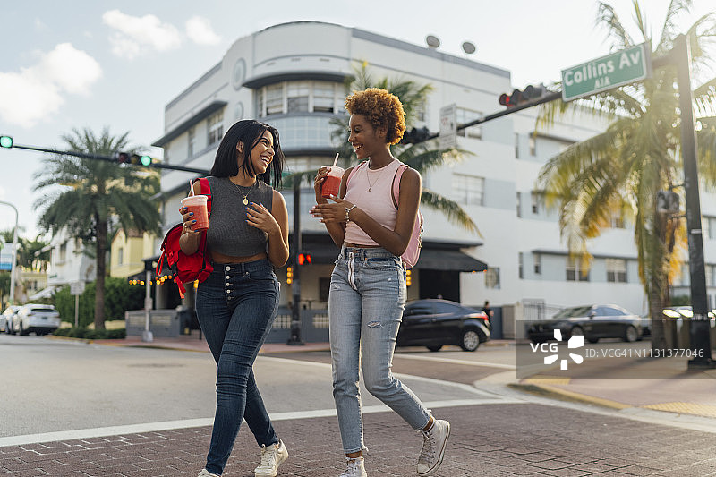 美国，佛罗里达，迈阿密海滩，两个快乐的女性朋友喝着软饮料过马路图片素材