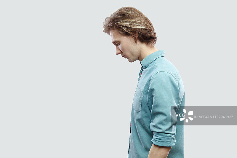 侧视图肖像悲伤英俊的长头发金发年轻人在蓝色休闲衬衫站着抱着头和感觉不好。图片素材