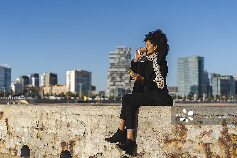 西班牙，巴塞罗那，一个拿着智能手机的女人坐在墙上喝着咖啡图片素材