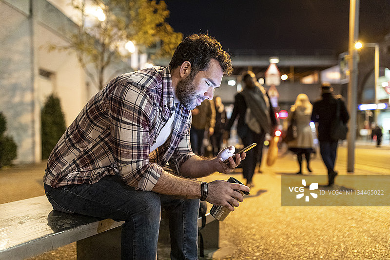英国，伦敦，一名微笑的男子坐在长凳上，看着他的手机图片素材