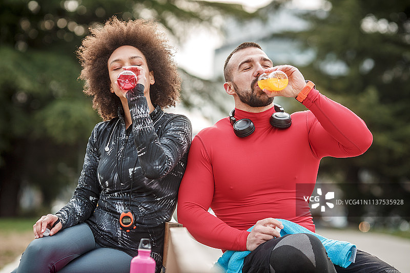 年轻的运动夫妇穿着运动服，并排坐在公园的长椅上，喝着能量饮料，一起度过美好的时光图片素材