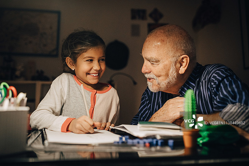 祖父和孙女坐在家里的桌子旁，微笑着互相看着图片素材