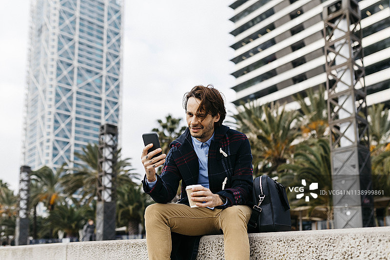西班牙，巴塞罗那，一个微笑的男人坐在城市里拿着外卖咖啡和手机图片素材