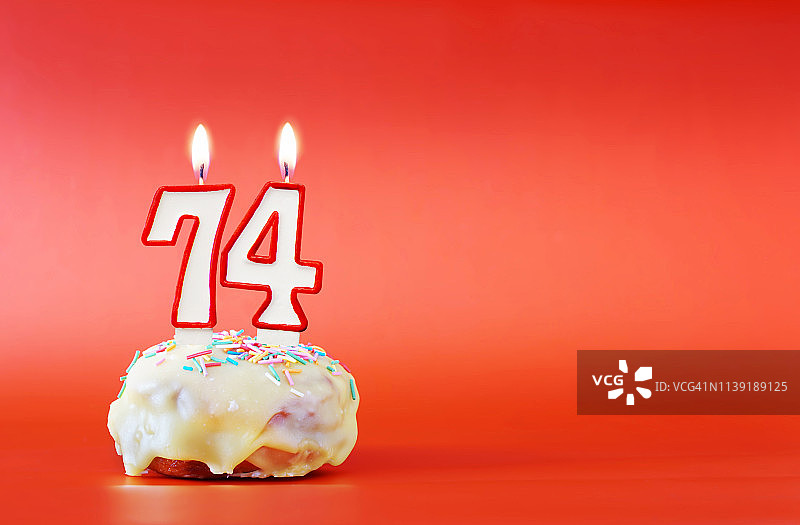 七十四岁生日。杯形蛋糕，上面有燃烧着的74号白色蜡烛。鲜艳的红色背景与复制空间图片素材