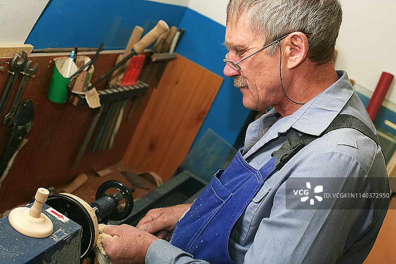 一位戴着眼镜、留着胡子的退休老人在车床上摆弄着一个木制工件。木匠的工作场所图片素材