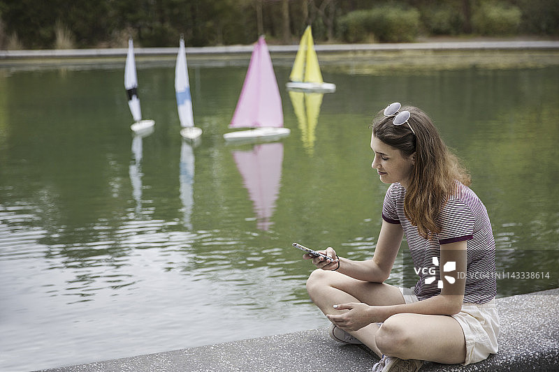 十几岁的女孩坐在公园湖边的挡土墙上使用智能手机图片素材