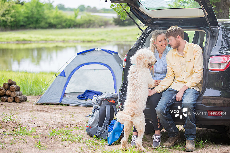 快乐的年轻夫妇和宠物狗坐在汽车附近的帐篷露营旅行图片素材