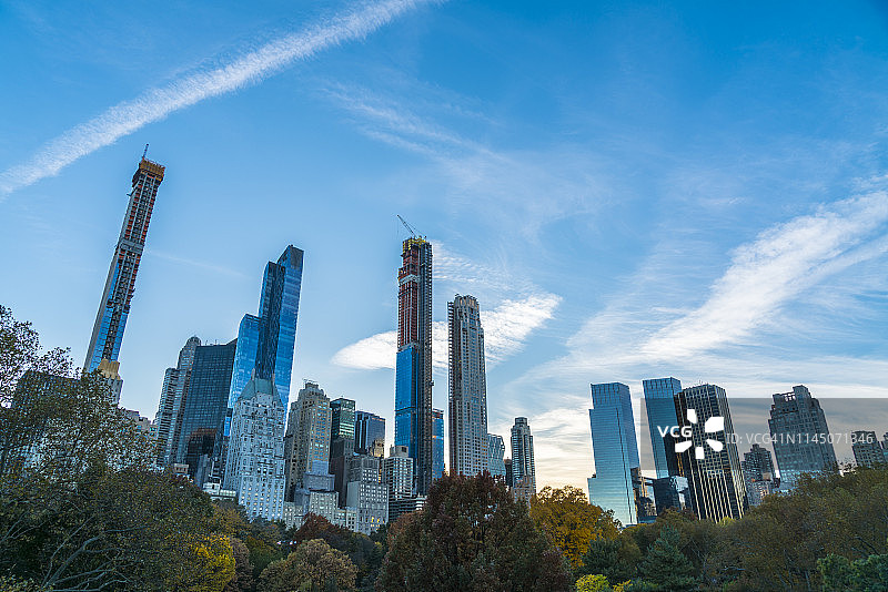 2018年11月4日，美国纽约曼哈顿中城摩天大楼在中央公园的树林中，飞机在秋日的蓝天上勾勒出凝结的痕迹。图片素材