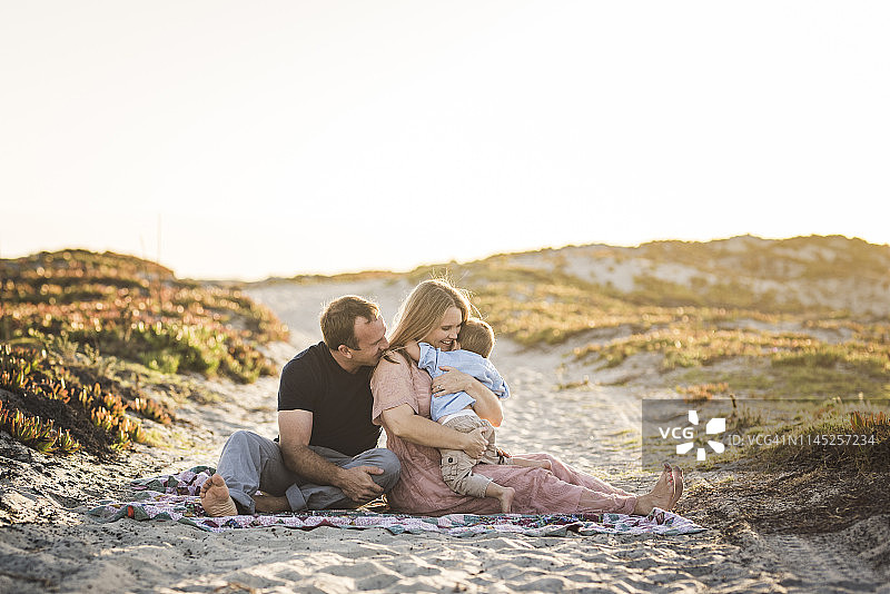 日落时分，晴朗的天空下，快乐的父母带着儿子坐在沙滩上图片素材