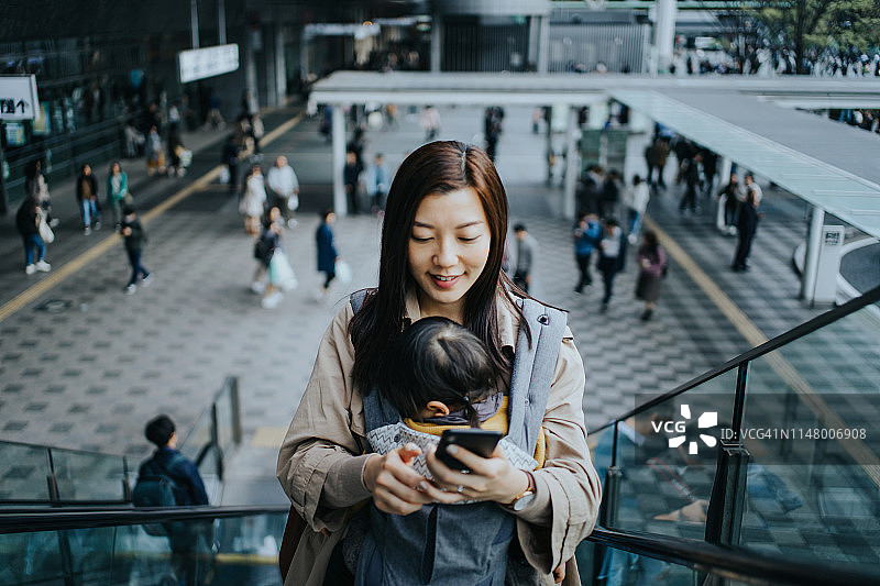 一位年轻的亚洲母亲带着小女儿在市中心坐自动扶梯时使用智能手机，背景是忙碌的上班族图片素材