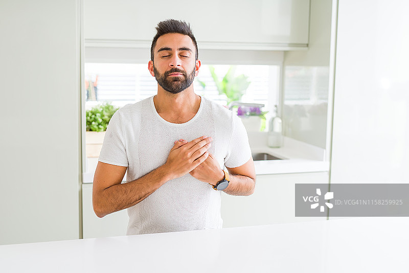 英俊的西班牙男子休闲白色t恤在家微笑与手在胸前闭上眼睛和感激的手势在脸上。健康的概念。图片素材