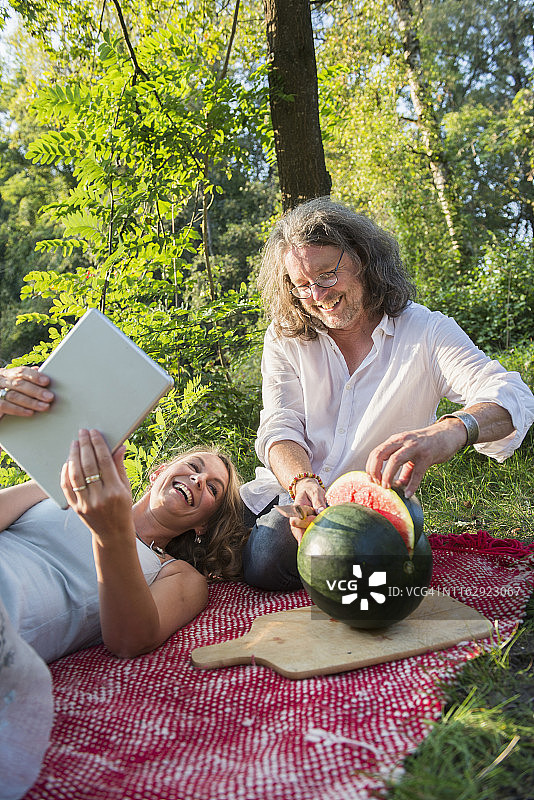 德国巴伐利亚，一对夫妇在野餐时用平板电脑切西瓜图片素材