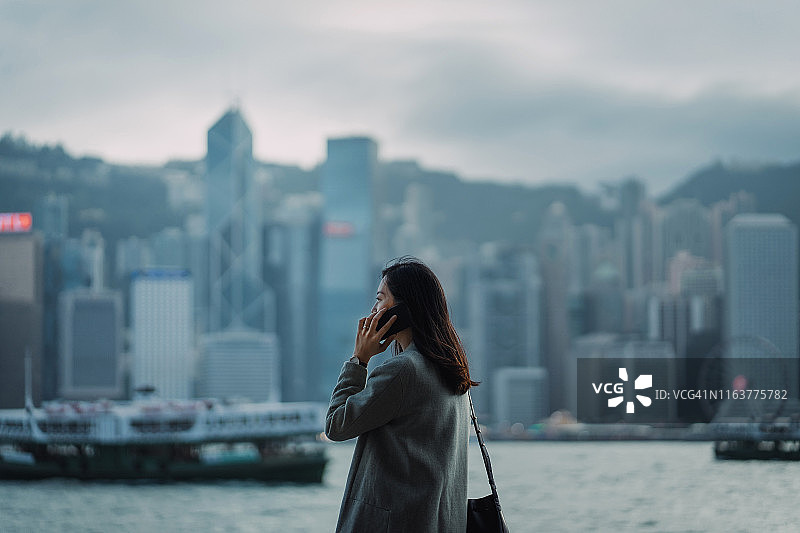 年轻的亚洲女商人在维多利亚港的海滨长廊上拿着智能手机，与香港市中心商业区的景色相映成趣图片素材