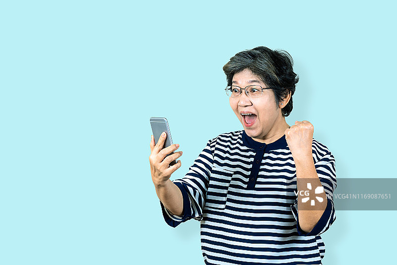 开朗的资深亚洲女人抱着和看智能手机在孤立的背景上感觉赢，庆祝和胜利。老年女性生活方式概念蓝色背景。图片素材