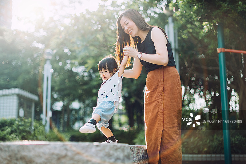 在一个阳光明媚的日子里，快乐的年轻亚洲母亲牵着小女儿的手，扶着她在石头上行走图片素材