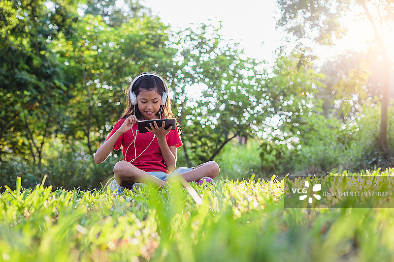 快乐可爱的亚洲小女孩坐在草地上，听音乐在户外的公园图片素材