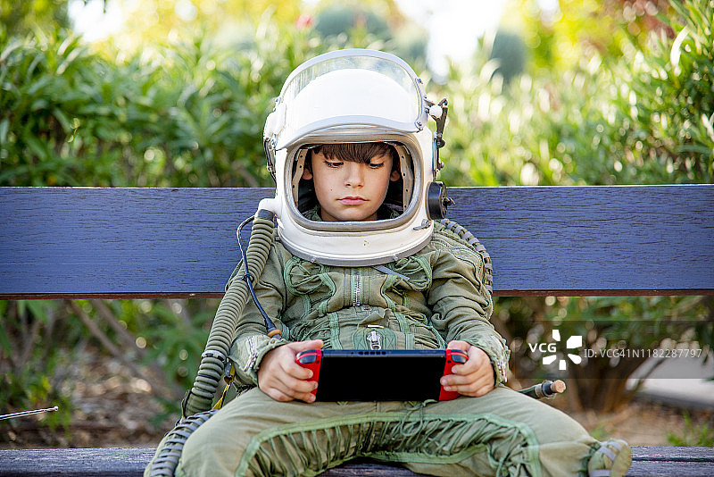 男孩穿着太空服，坐在长椅上，独自玩电子游戏图片素材