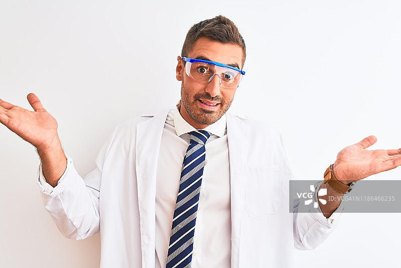年轻英俊的科学家男子戴着安全眼镜在孤立的背景下，愚蠢和困惑的表情与手臂和手举起。怀疑的概念。图片素材