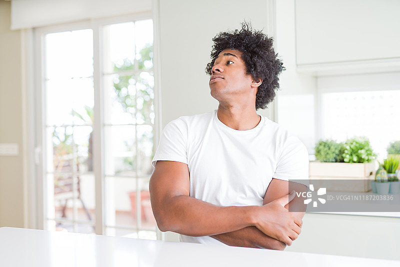 年轻的非洲裔美国男子穿着休闲的白色t恤坐在家里看着一边，双臂交叉，确信和自信图片素材