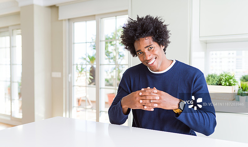 年轻的非洲裔美国男子穿着休闲毛衣坐在家里双手合十，十指交叉，微笑轻松愉快。成功和乐观图片素材