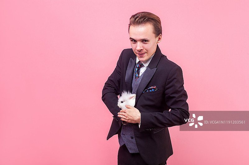 滑稽的正男把白兔藏在燕尾服里，魔术师在表演魔术。孤立在粉红色背景上图片素材
