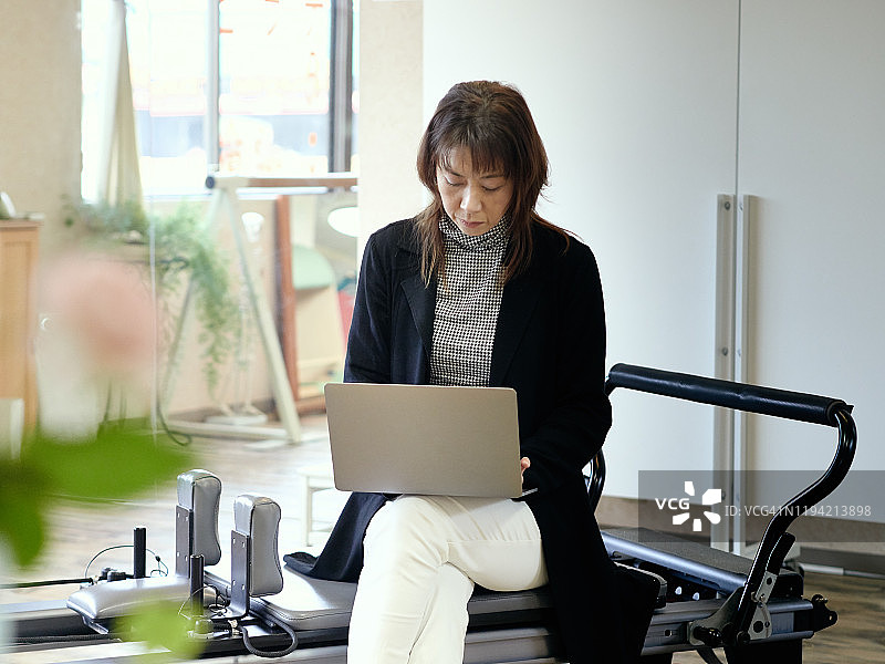 一名女教练坐在普拉提练习室的一台机器上使用笔记本电脑。图片素材
