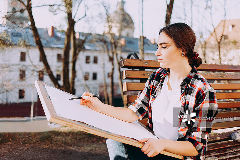 年轻的女艺术家坐在长凳上画画。画家艺术家手持画板和画笔图片素材