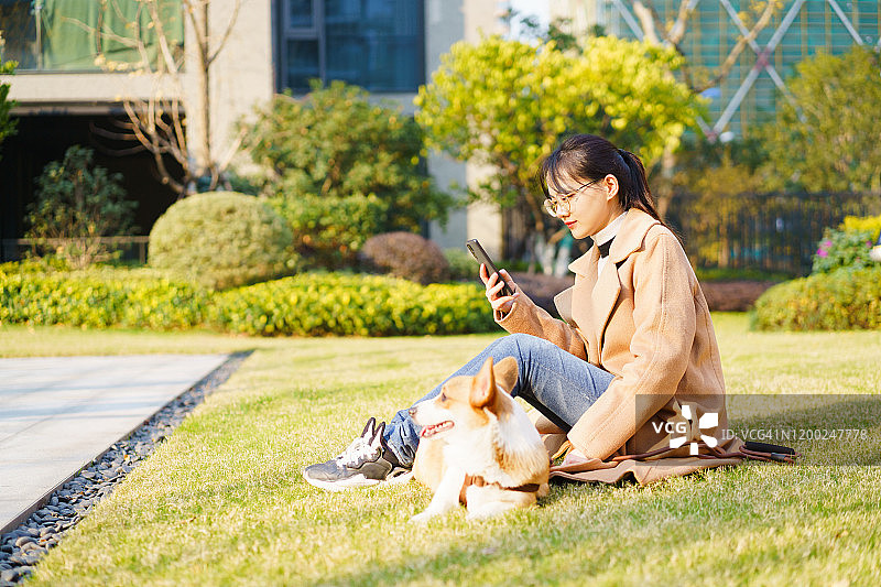 年轻的亚洲女性使用手机与柯基坐在草地上图片素材
