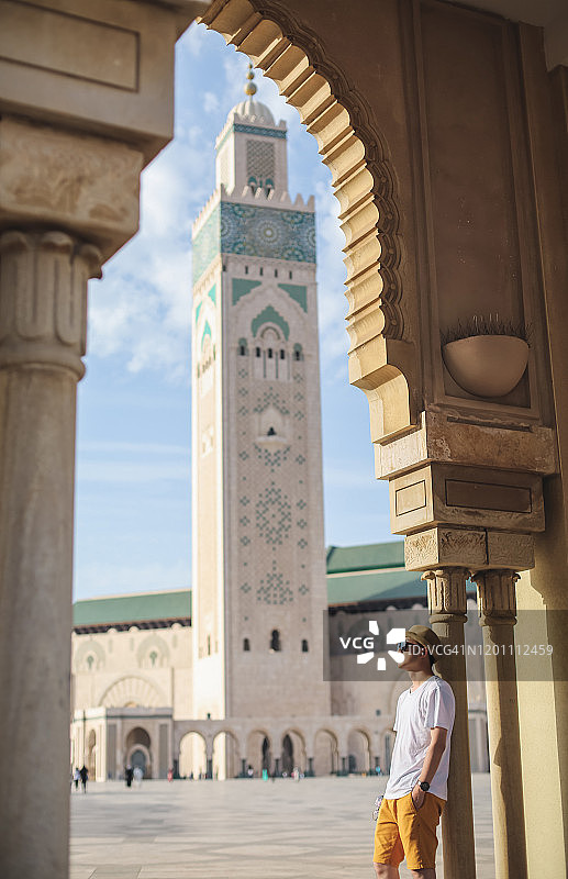 一名男子倚靠在哈桑二世清真寺的拱门和走廊上，清真寺尖塔是非洲摩洛哥卡萨布兰卡的地标图片素材
