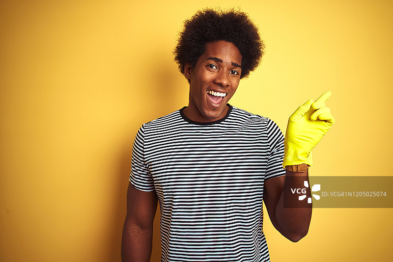 年轻的非洲裔美国人清洁用手套站在孤立的黄色背景非常高兴地用手和手指指向一边图片素材