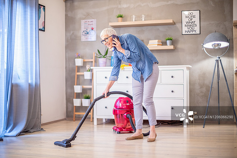 微笑的高级主妇用吸尘器打扫她的房子。图片素材