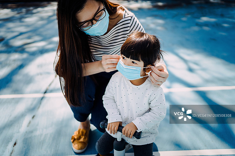 年轻母亲在操场上为小女儿戴上外科口罩，防止感冒、流感及病毒传播图片素材