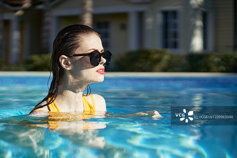 年轻女子在游泳池里摆姿势，穿着时髦的黄色泳衣和眼镜，湿的头发，干净的皮肤，阳光灿烂的一天图片素材