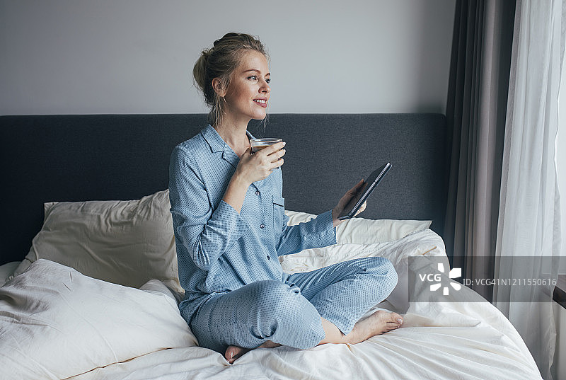 清晨惯例:一个快乐的金发女人坐在床上，在卧室的平板电脑上查看电子邮件的全身照图片素材