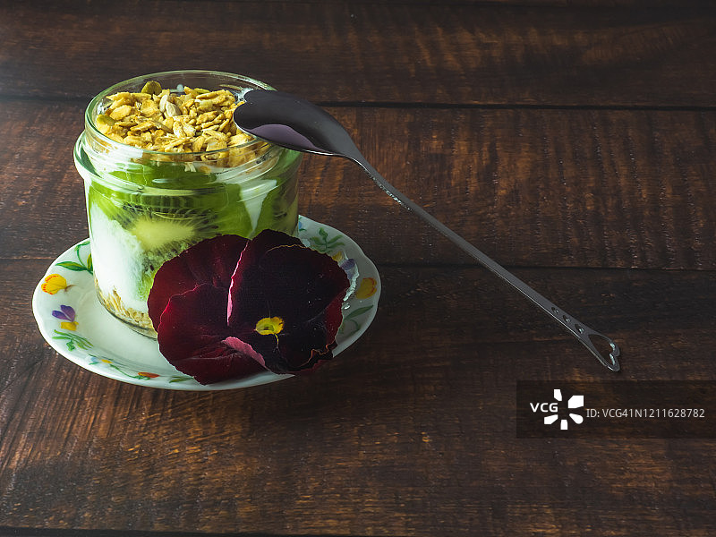 一杯含有猕猴桃和格兰诺拉麦片早餐的天然酸奶，在木桌上放一朵可食用的花和一把旧羊驼或银勺子图片素材