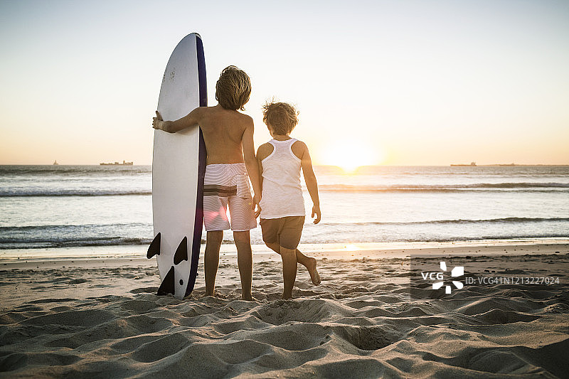 后视图的两个男孩与冲浪板站在海滩上日落图片素材